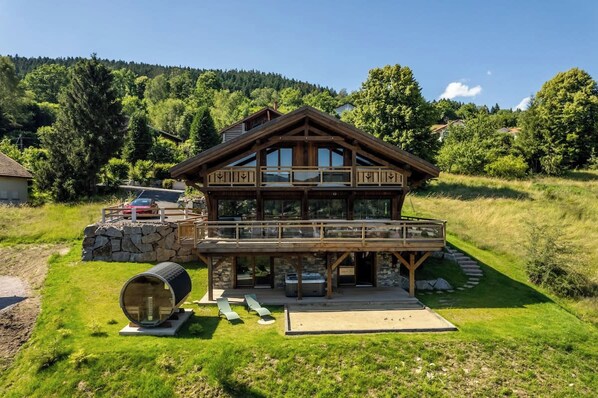 A wooden chalet with a balcony, a stone foundation, and a barrel in the foreground.