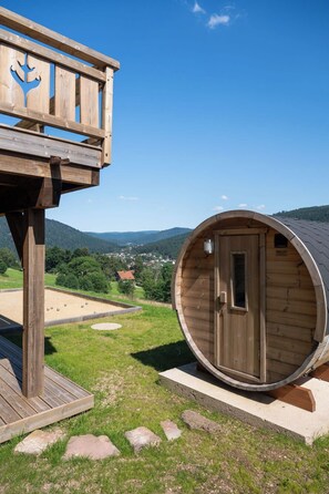 A wooden cabin with a circular door and a small window, situated on a grassy area with a view of a village in the background. - LE CERF - 12 pers -Sauna-Jacuzzi (Gérardmer)