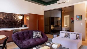 A modern living room with a purple sofa, a white sofa, and a coffee table with books. There is a kitchen area with a stove and a tiled backsplash.