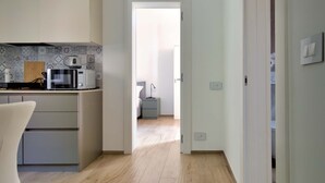 Una cocina moderna con microondas, tostadora y tabla de cortar de madera. Se ve un comedor con una silla. El ambiente tiene piso de madera clara y paredes blancas.