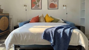 Ein Bett mit weißblauer Decke, einem blauen Kopfteil und bunten Kissen. Zwei Wandleuchten und ein abstraktes Kunstwerk sind an der Wand angebracht.