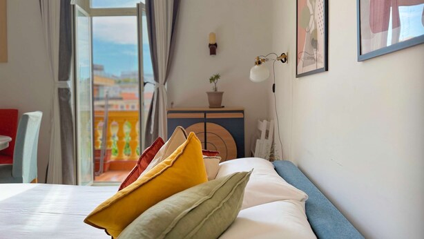 Ein Schlafzimmer mit Bett, Kissen, Nachttisch, Lampe und einem Fenster mit Vorhängen.