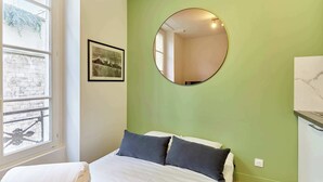 Een slaapkamer met een bed, een ronde spiegel, een ingelijst beeld en een raam met uitzicht op een gebouw.