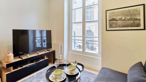 Een woonkamer met een grote flatscreen televisie, een houten tv-meubel, een eettafel met twee kommen en wijnkaraffen, en een ingelijste foto aan de muur.