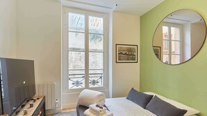 Een hotelkamer met een bed, een televisie, een raam met uitzicht op gebouwen en een ronde spiegel aan de muur.