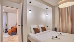 Une chambre d’hôtel moderne avec un grand lit, deux lampes suspendues, une table de chevet et une vue sur le couloir.