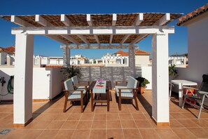 A covered patio with a table and chairs, a pergola, and a view of buildings in the background. - El Recreo 10 (Torrox Costa)