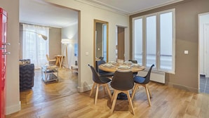 Een eetruimte met een ronde tafel voor vier personen, omringd door stoelen. Er is een groot raam met meerdere ruiten, een vloerlamp en een spiegel aan de muur.