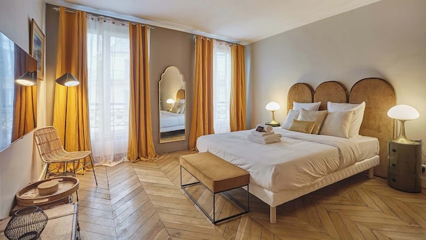 Een hotelkamer met een groot bed, een bankje, een stoel, een spiegel en een raam met gordijnen.