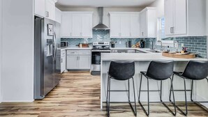 A modern kitchen with white cabinets, a stainless steel refrigerator, a central island with bar stools, and a blue-tiled backsplash. - Nautical Nest (Rockport)