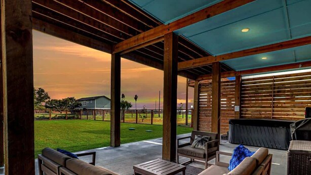 An outdoor patio area with wooden furniture, a table, and a view of a grassy field and buildings in the distance. - Nautical Nest (Rockport)