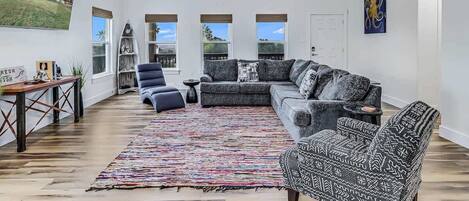 A spacious living room with a large sectional sofa, a ceiling fan, and a view of a house through large windows.