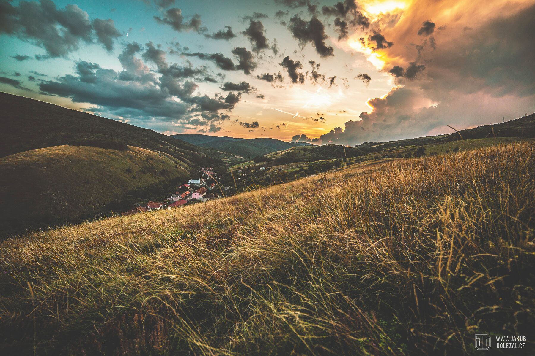A rural landscape with rolling hills, a small village, and a sunset.