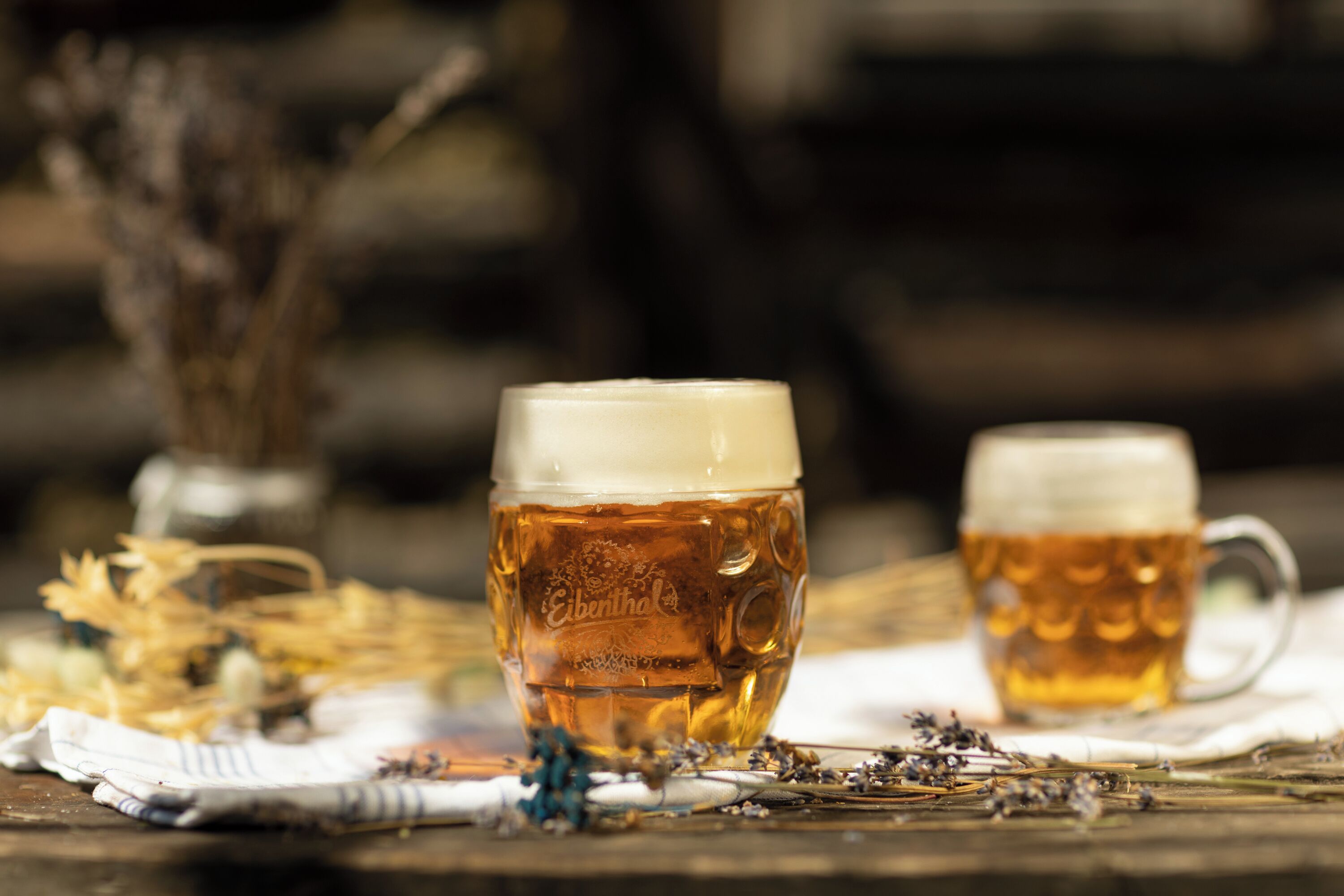 Deux verres de bière posés sur une surface en bois, avec un tissu et des fleurs séchées.