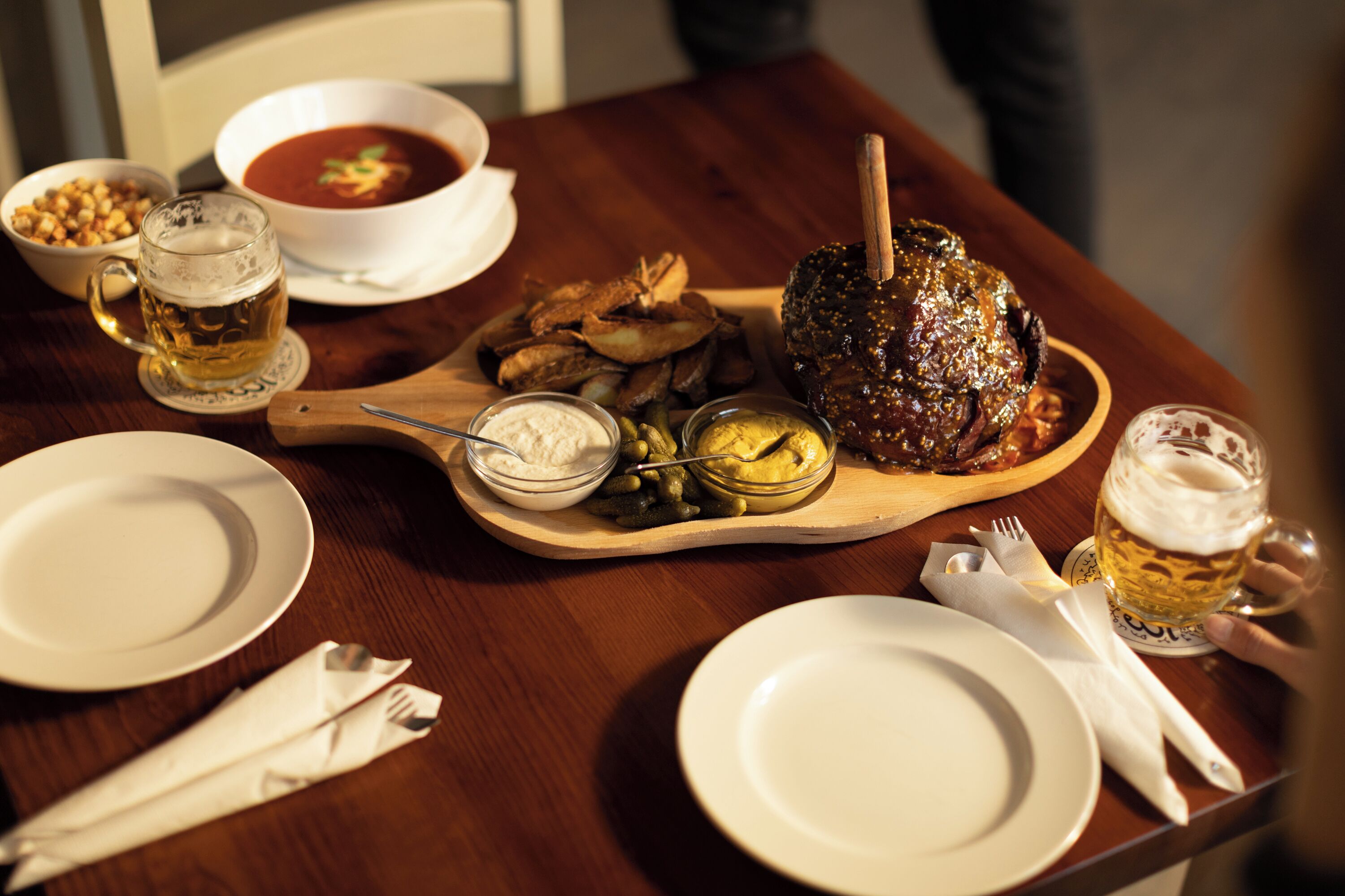 A wooden table with a roasted meat centerpiece, bowls of soup and condiments, and glasses of beer.