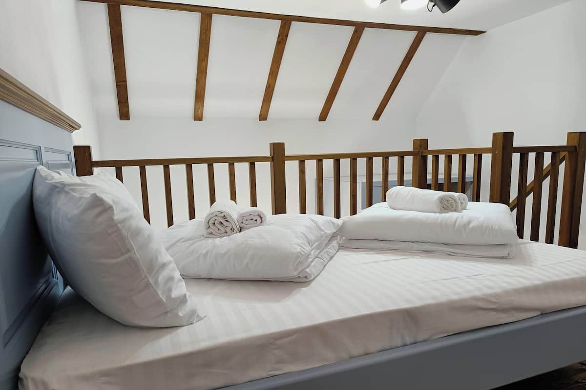 A neatly made bed with white linens and rolled towels, a wooden headboard, and a sloped ceiling with exposed beams.