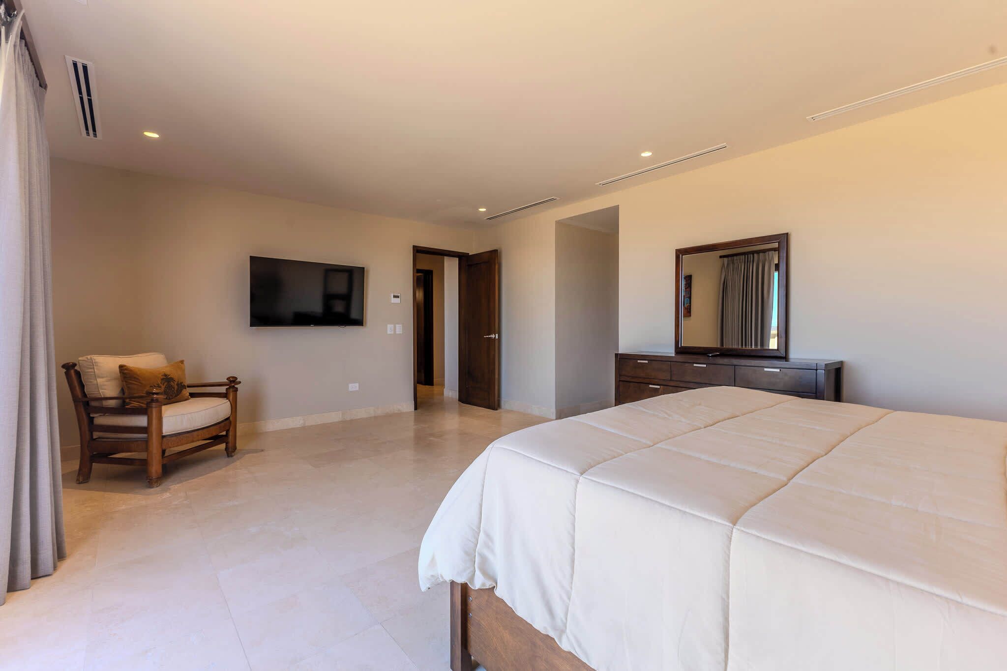 A bedroom with a large bed, a wooden chair, a TV mounted on the wall, and a dresser.