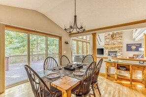 A spacious dining area with a large wooden table, chairs, and a chandelier. There is a stone fireplace and a television mounted on the wall. - Brightside Retreat - Resort Pool, Games, Pets (Mount Hood Village)