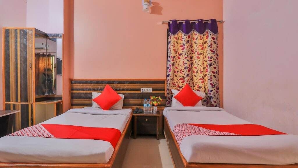 Une chambre d’hôtel avec deux lits simples, chacun agrémenté d’un oreiller rouge et d’une couvre-lit à motifs. On y trouve une tête de lit en bois, une table de chevet avec une lampe et un miroir. Les murs de la chambre sont peints en rose et une fenêtre est ornée de rideaux à motifs.