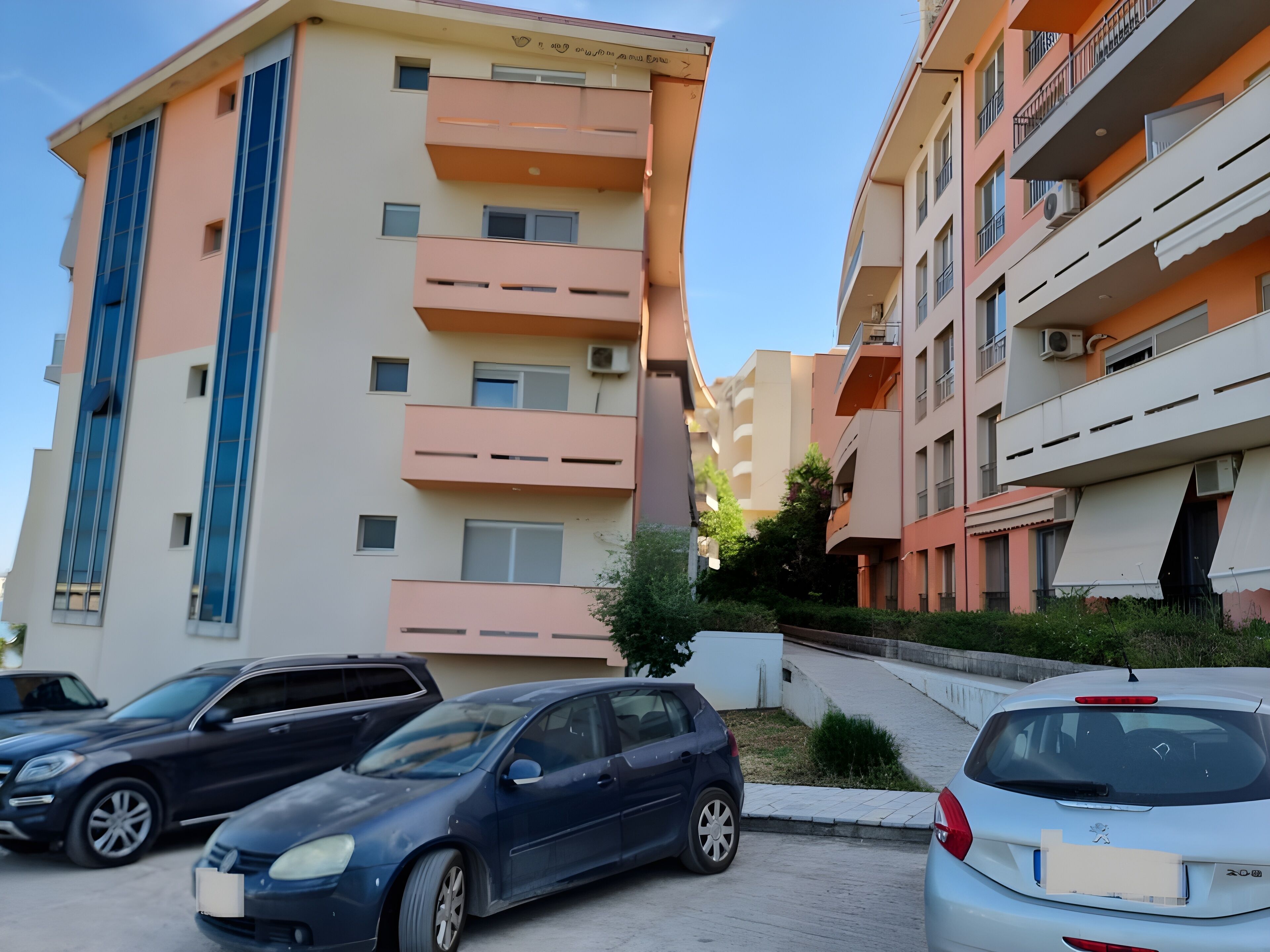 A multi-story residential building with balconies and a parking area with several cars.