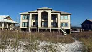 Een groot, twee verdiepingen tellend huis met een balkon en een overdekte veranda, omringd door zand en hoge grassen.