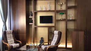 Un salon moderne comprenant un canapé, deux fauteuils, une table basse, un téléviseur fixé à un mur en bois, ainsi que des objets décoratifs tels qu’un trophée et des figurines.