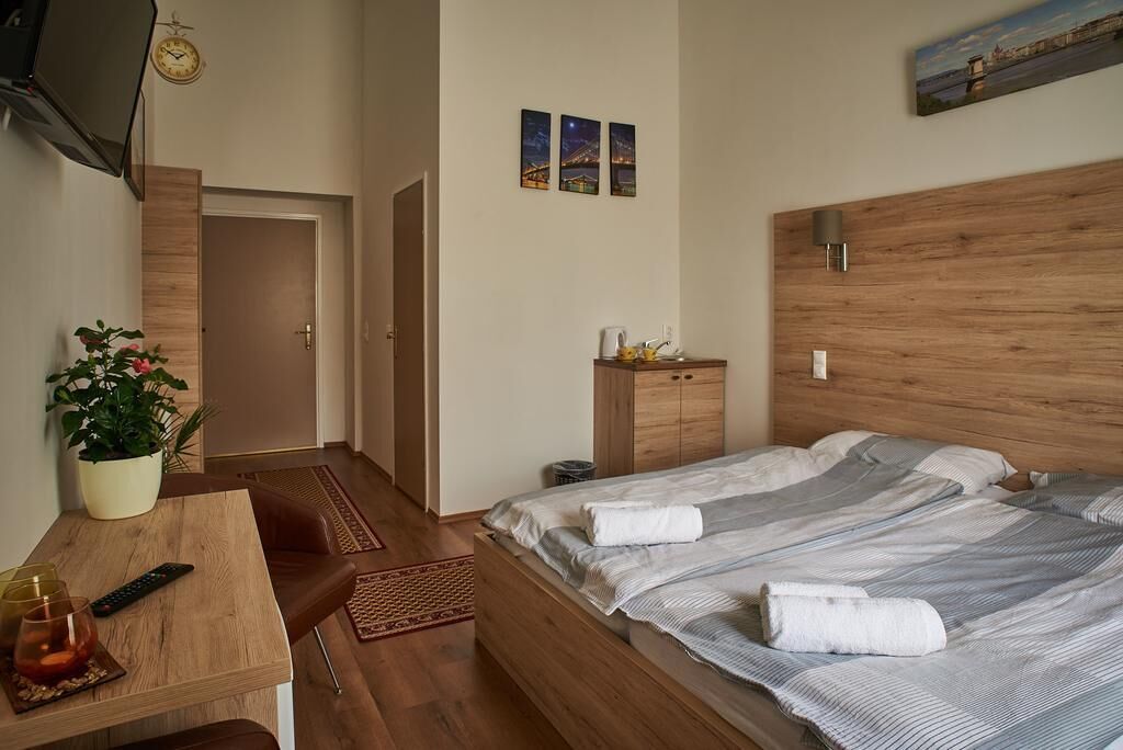 Une chambre avec une tête de lit en bois, un lit avec deux oreillers, une table de chevet avec une télécommande, une plante et une horloge au mur.