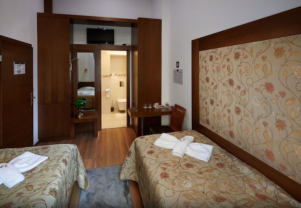 A hotel room with two beds, a desk, a chair, a television, and a bathroom visible through an open door. - Vinci Apartman Szombathely (Szombathely)