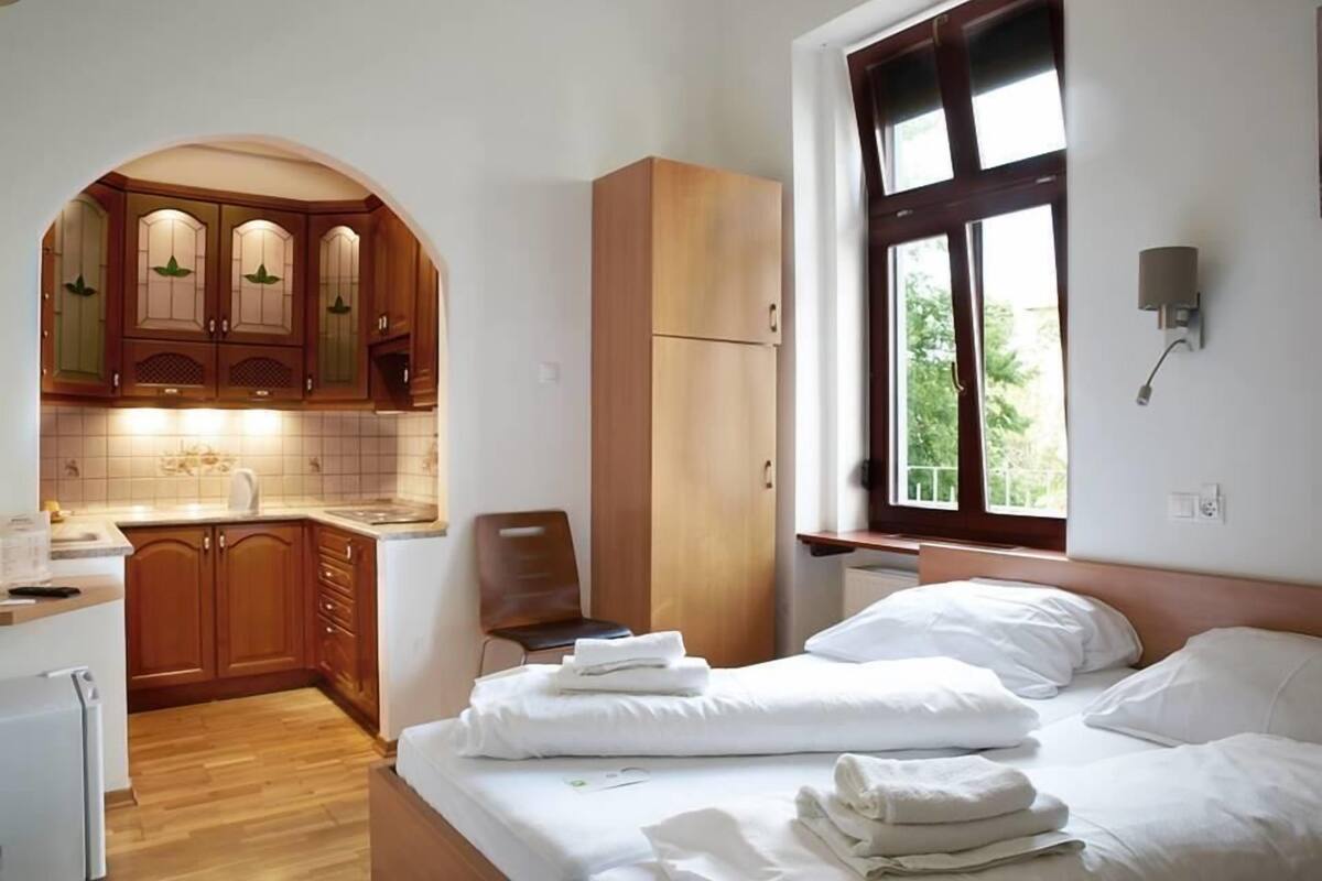 A hotel room with a kitchen area, a bed with white linens, and a window with a view of greenery.