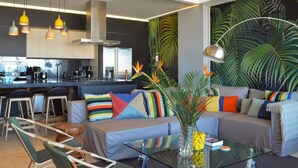 Una sala de estar moderna con un sofá, cojines coloridos, una mesa de centro de vidrio y una cocina al fondo.