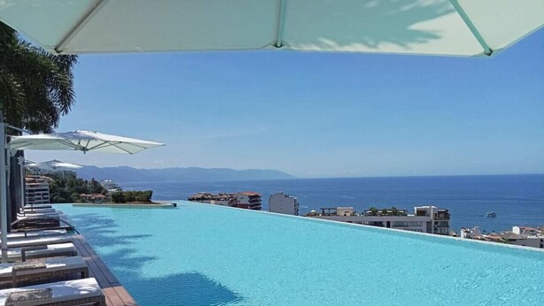 Una piscina infinita de color azul cristalino con sillas de descanso y sombrillas, con vistas a una ciudad costera.