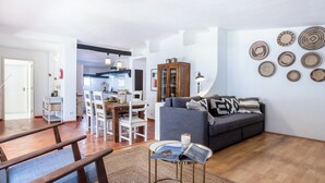 Een moderne woonkamer met een grijze bank, een houten eettafel en stoelen. In de achtergrond bevindt zich een keuken met witte kasten en een hangende lamp.