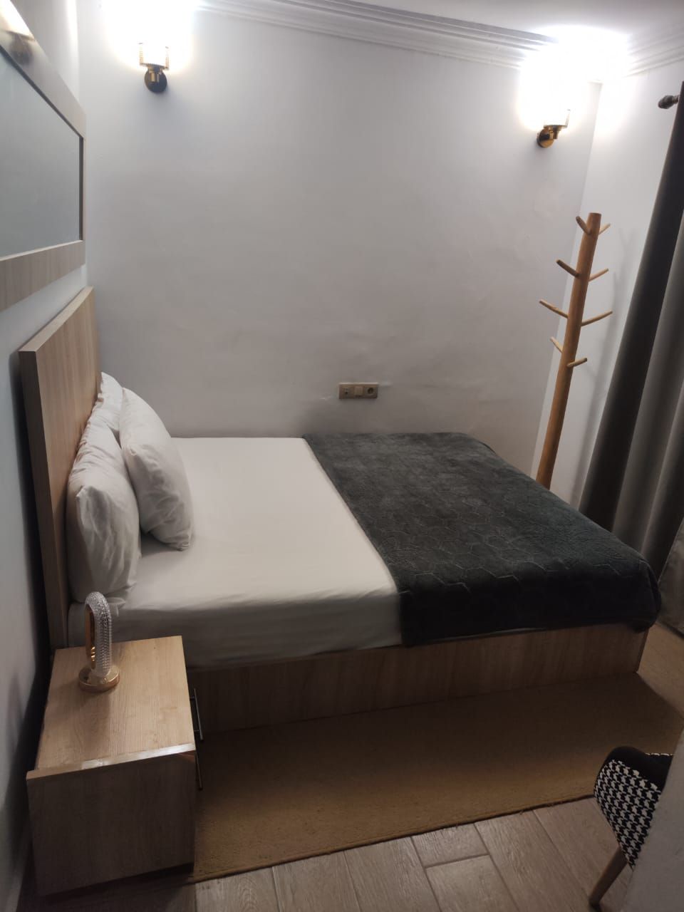 A small, neatly arranged bedroom with a single bed, a nightstand, a lamp, and a coat rack.