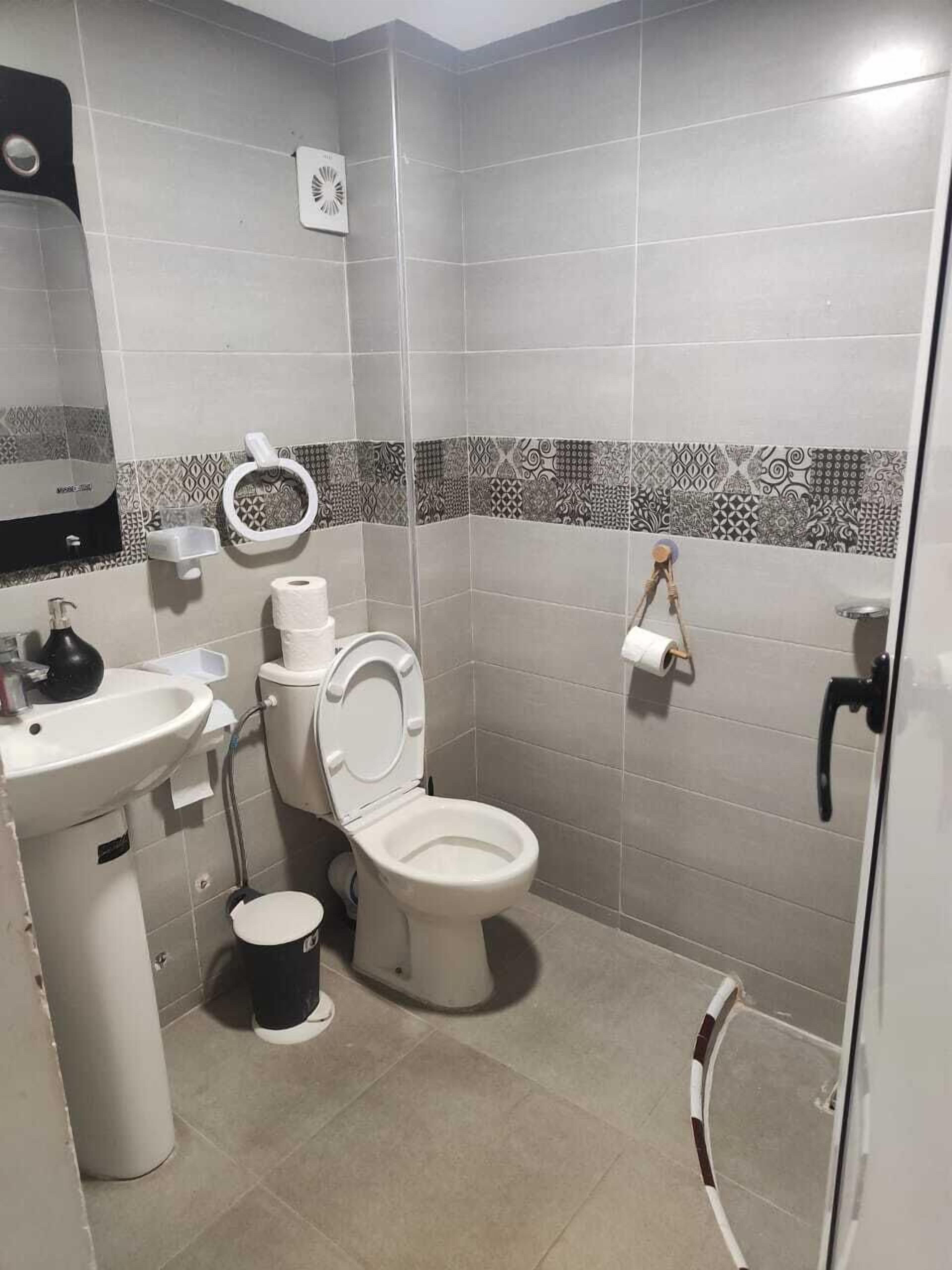 Une salle de bain comprenant des toilettes, un lavabo et un bidet. On y trouve un porte-rouleau de papier toilette et une poubelle.
