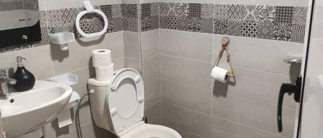 Une salle de bain comprenant des toilettes, un lavabo et un bidet. On y trouve un porte-rouleau de papier toilette et une poubelle.