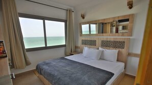A hotel room with a large bed, a view of the sea through a window, a wooden headboard, and a small shelf with books and a clock. - HOTEL PLAYA DAKHLA (Dakhla)