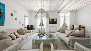 A living room with a white ceiling, a glass coffee table, two sofas, a wicker chair, a television, and a painting on the wall. - Villa Vinea Paros (Paros)