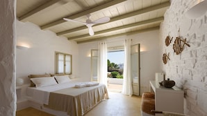 A bedroom with a bed, a ceiling fan, a desk with a lamp, and a view of the outdoors. - Villa Vinea Paros (Paros)