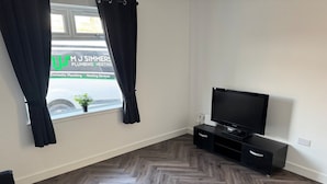 A modern living room with a herringbone wood floor, a black sofa, a black ottoman, a flat-screen TV on a black stand, and a window with black curtains.