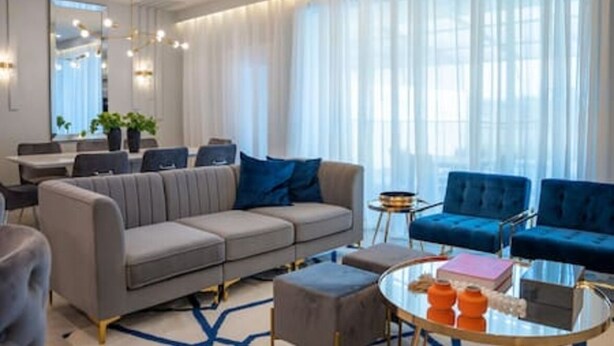 Una sala de estar moderna con un sofá gris, sillones azules, una mesa de centro de vidrio y un comedor al fondo.