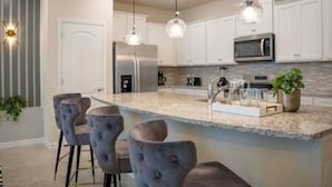 Una cocina moderna con gabinetes blancos, un refrigerador de acero inoxidable, un microondas y una encimera de granito. Hay tres taburetes de bar y lámparas colgantes sobre la encimera.