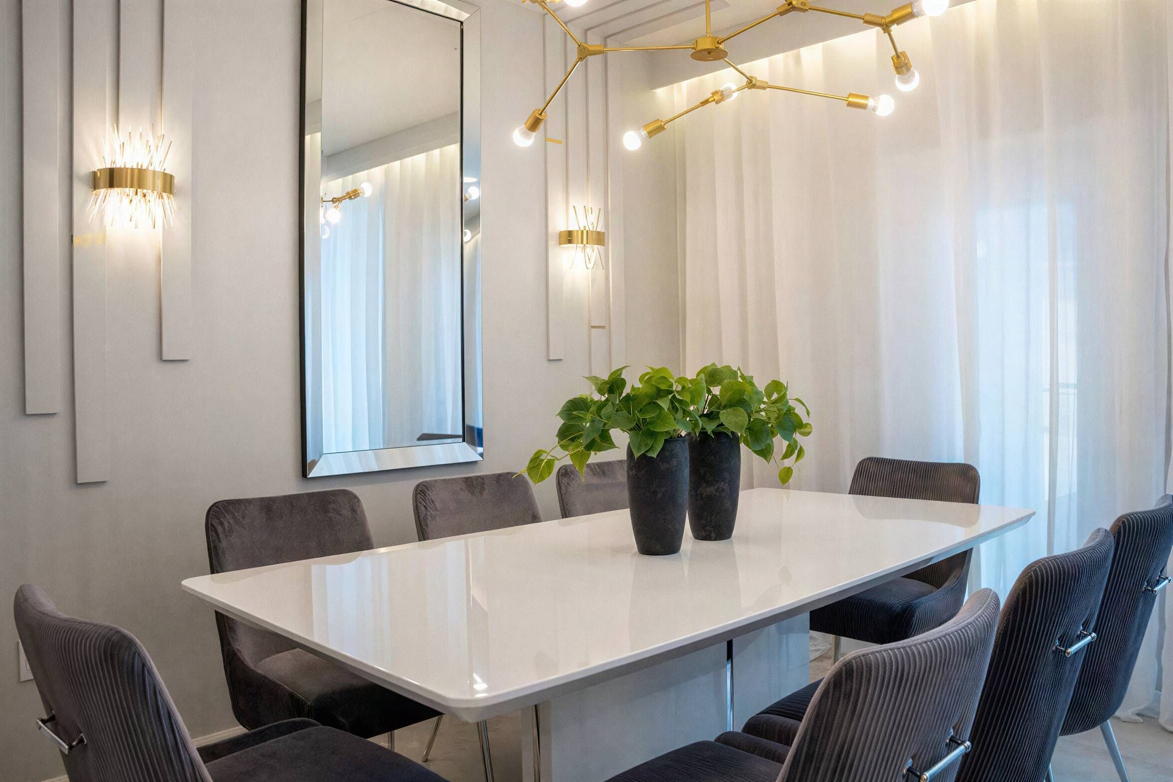 Un salón de conferencias con una mesa blanca, seis sillas, dos plantas en macetas, un espejo grande e iluminación instalada en la pared.