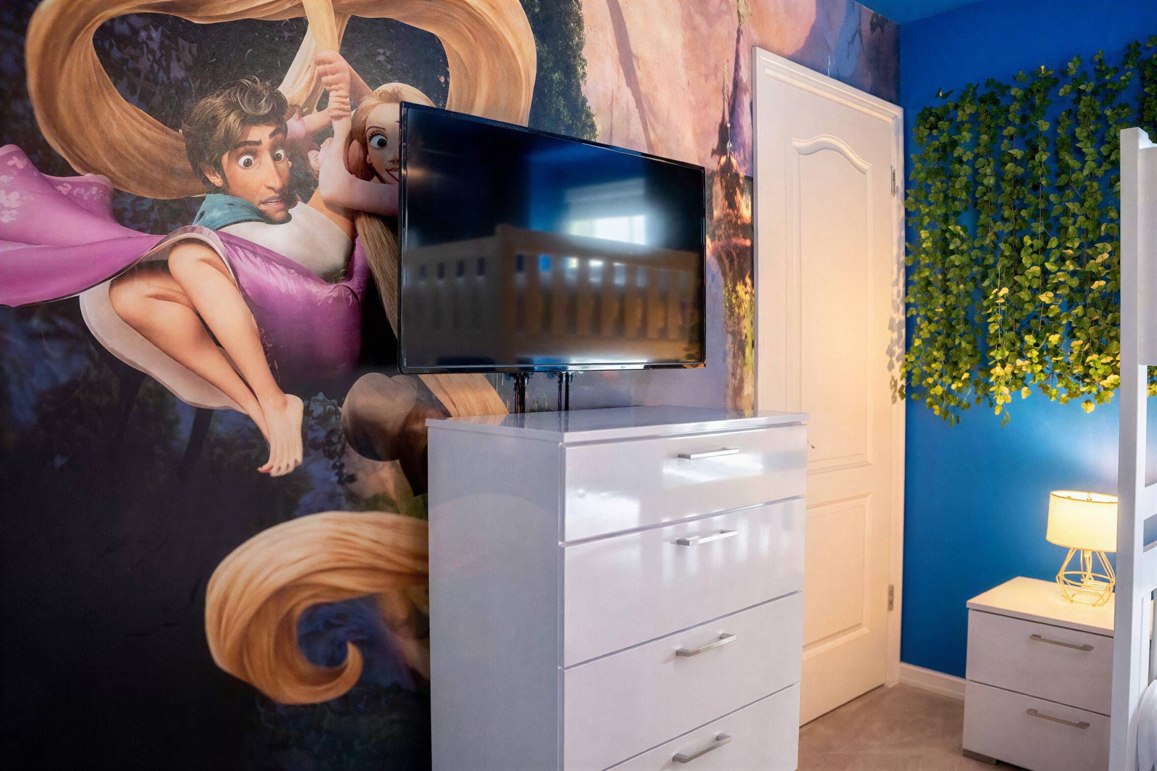 Una habitación con una televisión sobre un tocador, un mural en la pared y un muro azul con follaje verde.