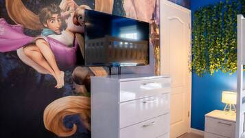 Una habitación con una televisión sobre un tocador, un mural en la pared y un muro azul con follaje verde.