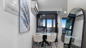 A modern hotel room with a large mirror, a dining area with a glass table, and a view of the cityscape through large windows.