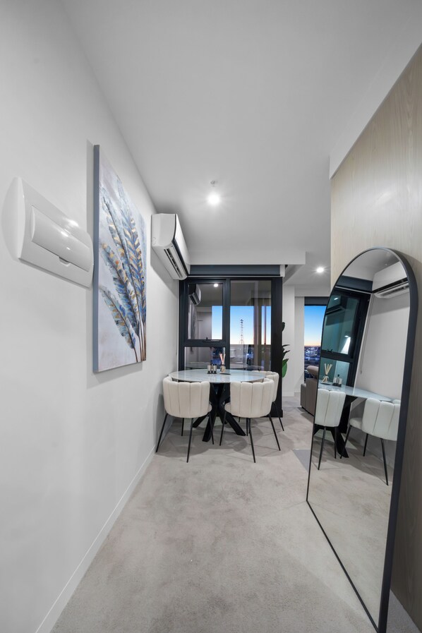 A modern hotel room with a large mirror, a dining area with a glass table, and a view of the cityscape through large windows.