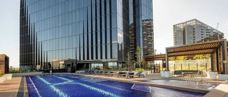 A modern building with a reflective glass facade, a rooftop pool with blue water, and outdoor seating areas.