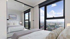 A modern hotel room with a large bed, a seating area, and a city view through large windows.