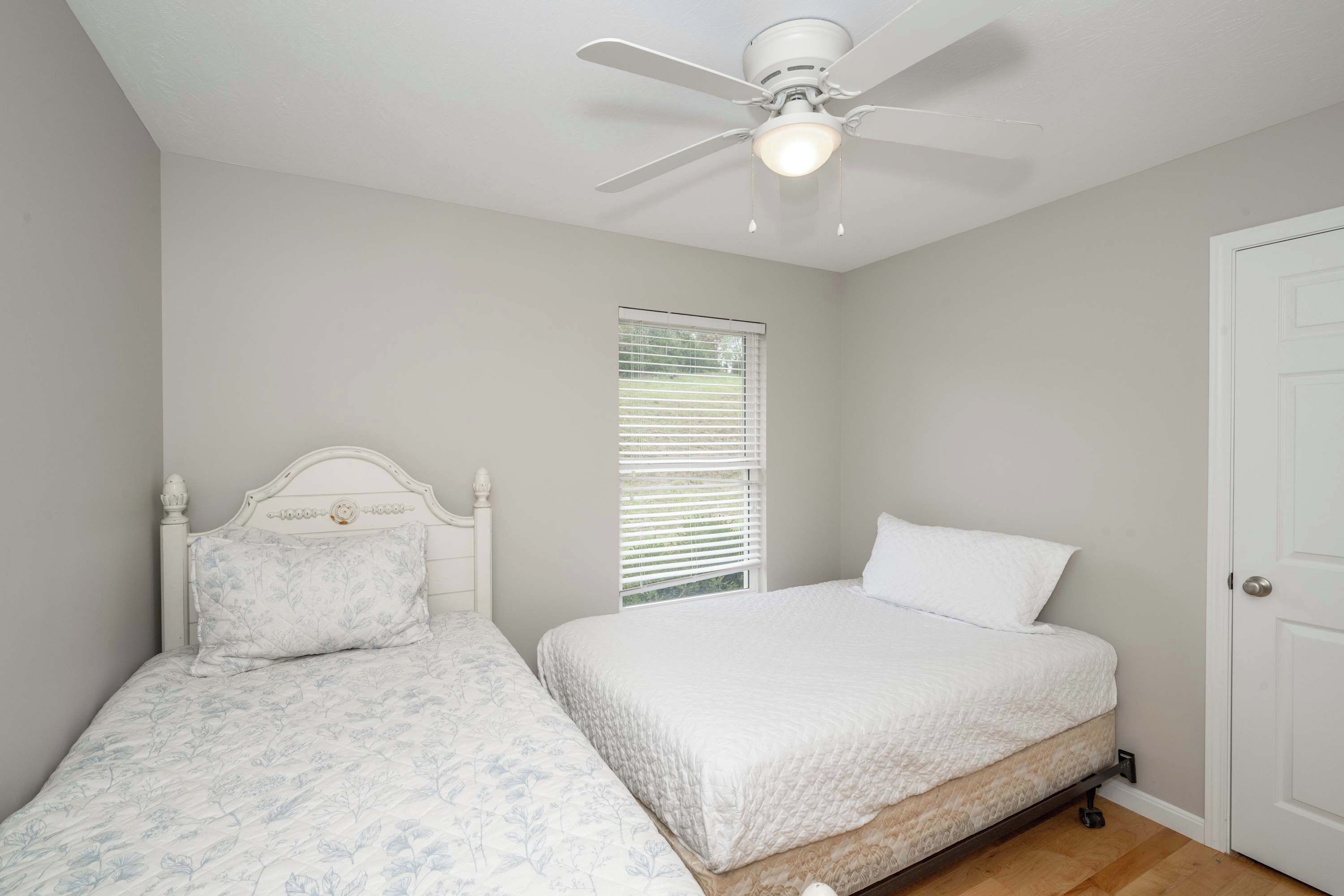 A bedroom with two beds, a ceiling fan, a window with blinds, and a white door.