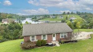 A single-story brick house with a front porch, a small yard, and a view of a river and hills in the background.